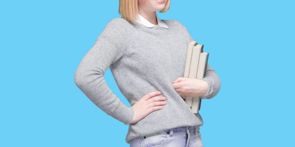 Young woman in gray sweater holding books against a blue background.