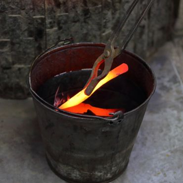 Tongs holding a glowing hot metal piece over a bucket of fire.