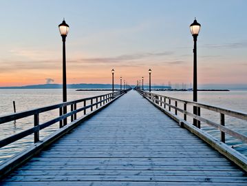 Long wooden pier with lampposts extending into calm water at sunset.
