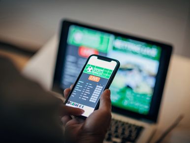 Person using a phone to place live sports bets with a laptop in the background.