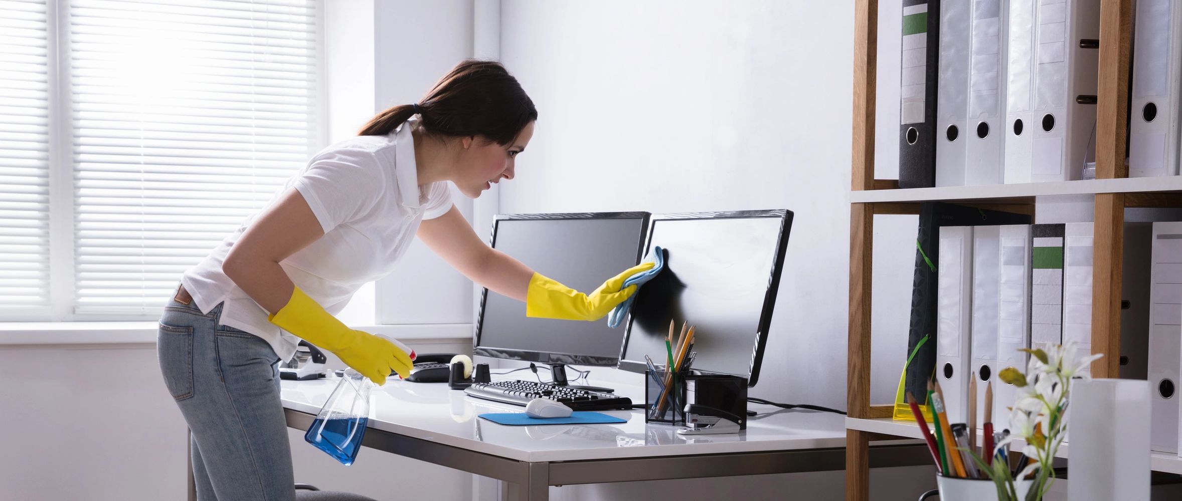 Woman wiping computer monitor, while wearing gloves, in an office setting. 