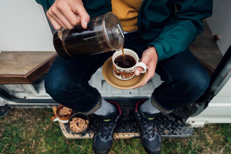 A directly above shot of a young man sitting on the side of his motor home pouring a cup of coffee.