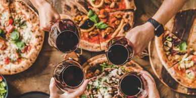 Friends toast with red wine over a table full of assorted pizzas.