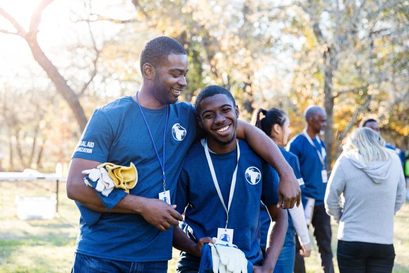 At the community park cleanup, the older brother smiles at and puts his arm around his younger brother.