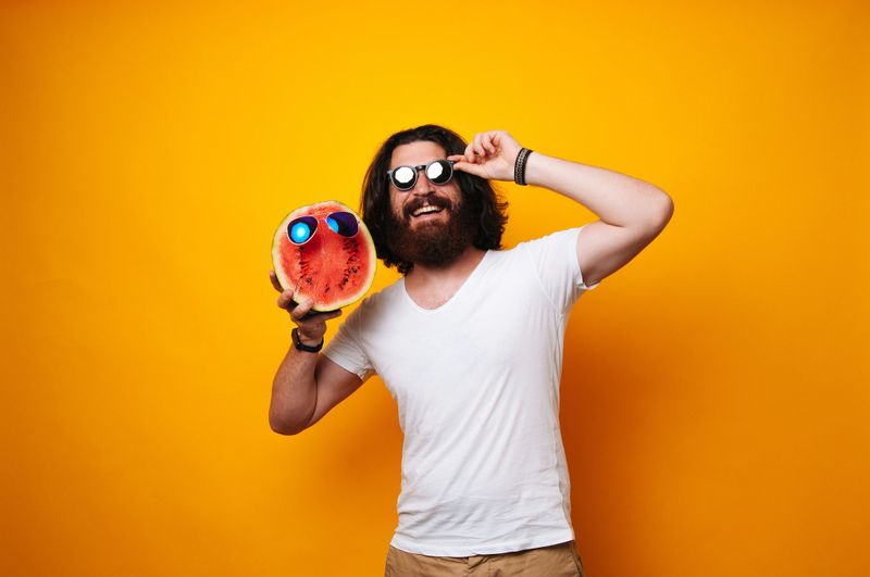 Half of Watermelon face with glasses held by a bearded man. Hispter wearing white shirt with long hair. Summer offer, vacation, holiday rest. Traveling and tourism concept. Summer buddy. Vegetarian or Vegan concept.