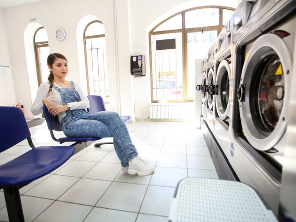 A woman waits in a laundromat, sitting on a blue chair.