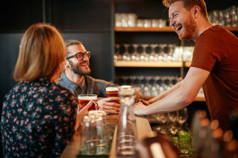 Cheerful friends leaning on bar counter, drinking beer and chatting with bartender. Night out.