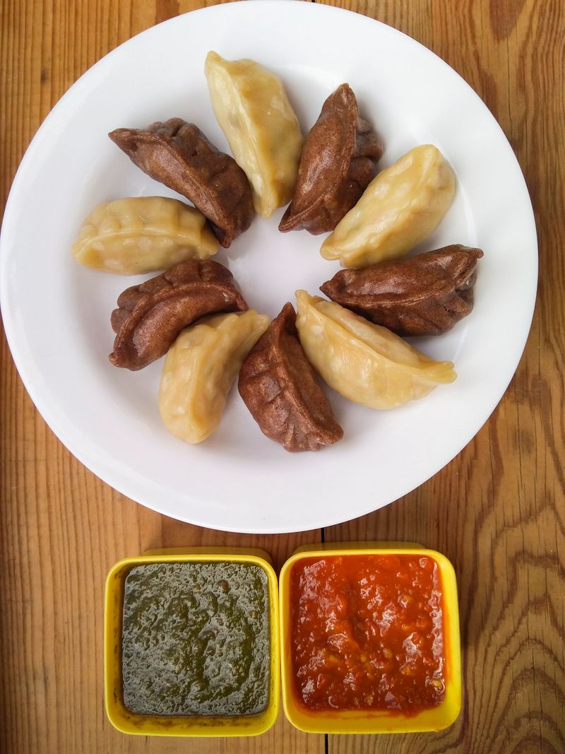 Two different types of momos (dumpling) with red and green chutney in white plate on wooden table.