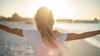 Person with arms outstretched facing the sun on the beach.