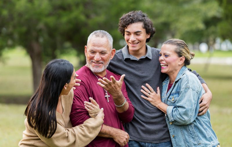 A happy, affectionate, Hispanic family standing together in the park, side by side, smiling and conversing. Father and son, a 16 year old teenage boy, are in the middle, between the mother and adult daughter, in her 30s.