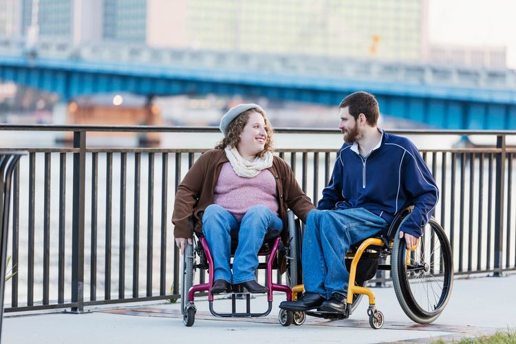 A smiling couple in wheelchairs holding hands outdoors by a river and bridge.
