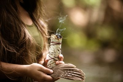 A woman holding a smoldering sage bundle and feathers outdoors.