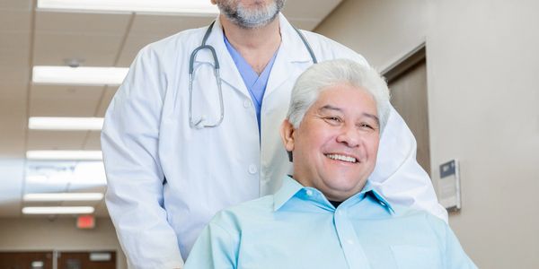 Smiling doctor standing behind a happy elderly patient in a wheelchair in a hospital corridor.