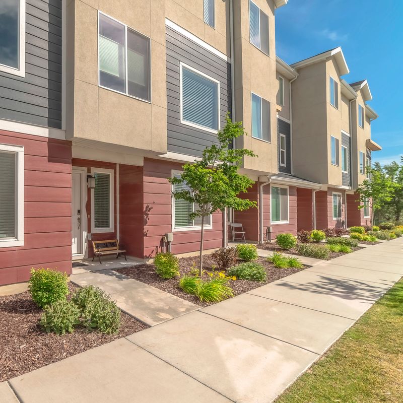 Square frame Townhomes exterior with red gray and beige wall against blue sky on a sunny day. Concrete pathways on the yards lead to the white front doors.