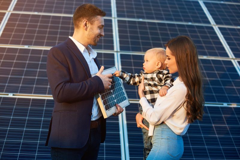 Cheerful kid in his mother arms examines the solar battery that dad holds, against background solar panels. Concept of family warmth and comfort in modern world with continuously advancing technology