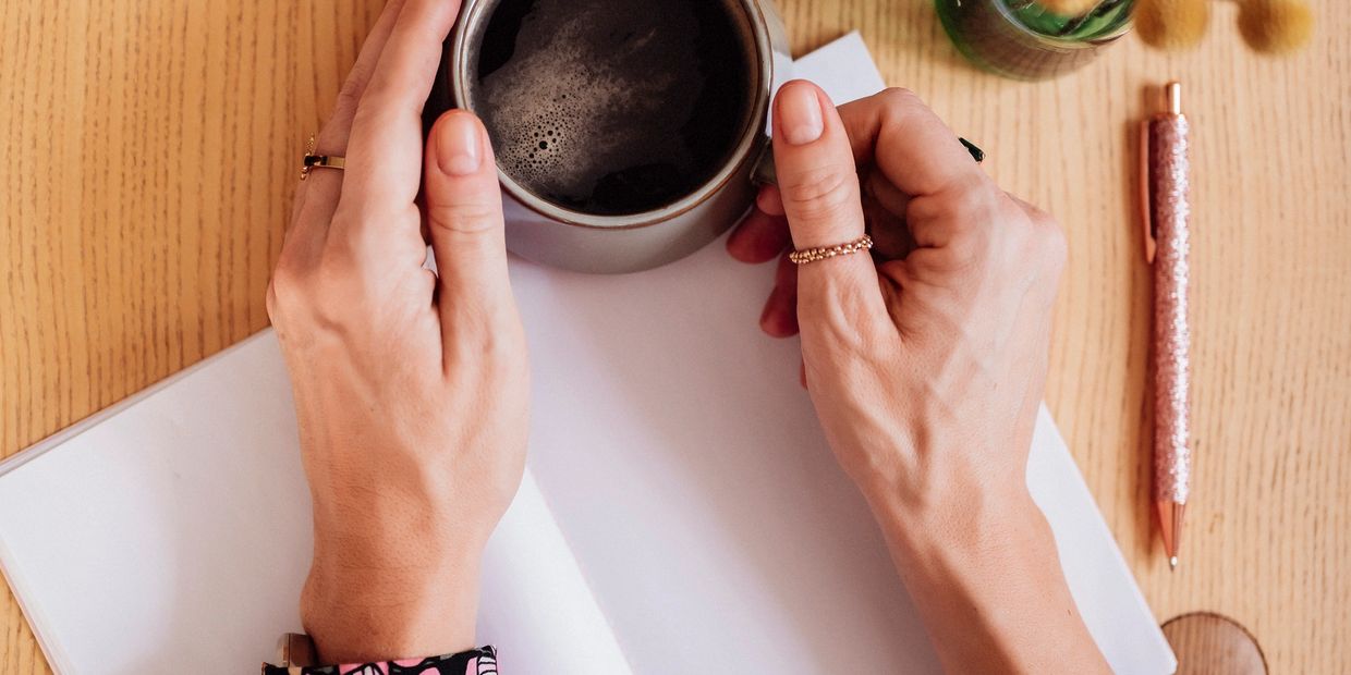 Hands holding a cup of coffee over an open notebook on a wooden table.