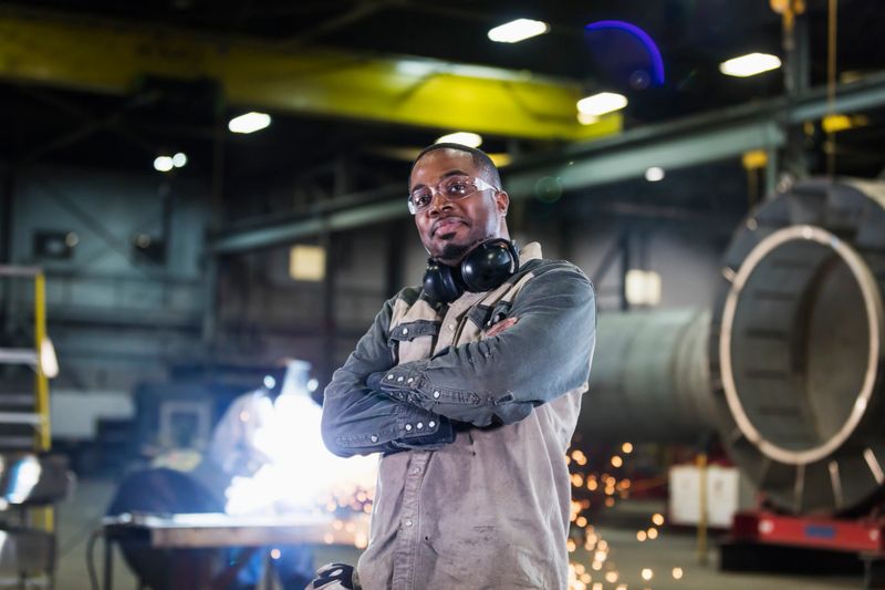A mid adult African-American man in his 30s working in a metal fabrication factory. He is standing in the foreground with his arms crossed, looking at the camera. Sparks are flying in the background where a worker is welding.