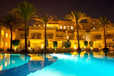 Luxury hotel illuminated at night with pool and palm trees.