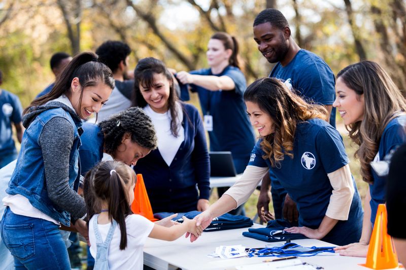 While other people register, a mid adult volunteer reaches across the table to shake hands with a young girl.  Other volunteers smile and watch.