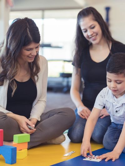 Young boy playing with puzzle pieces while two women watch and smile.