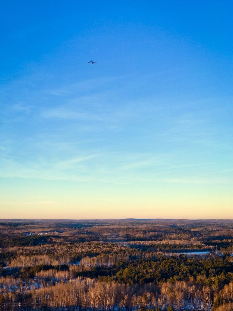 Mountaintop view, taken in Iron Mountain, Michigan in The Upper Peninsula of Michigan. The photograph has high up views of miles of different colored trees and a big view of a multicolored sunset or sunrise sky filled with blues, oranges, and pinks. There is a small airplane flying in the sky.