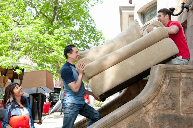 two male friends carrying couch up stairs to new home, woman carrying lamp and pillow, moving truck in the background, urban environment