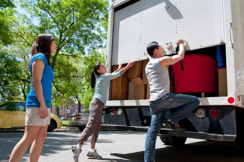group of friends standing at the back of small moving truck, lifting up rear door, truck filled with moving boxes and furniture, urban environment