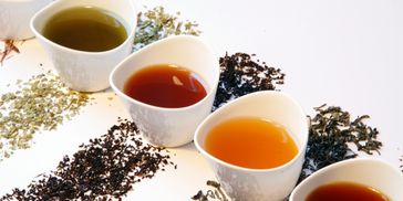 Five heart-shaped cups of tea with corresponding loose tea leaves on white.