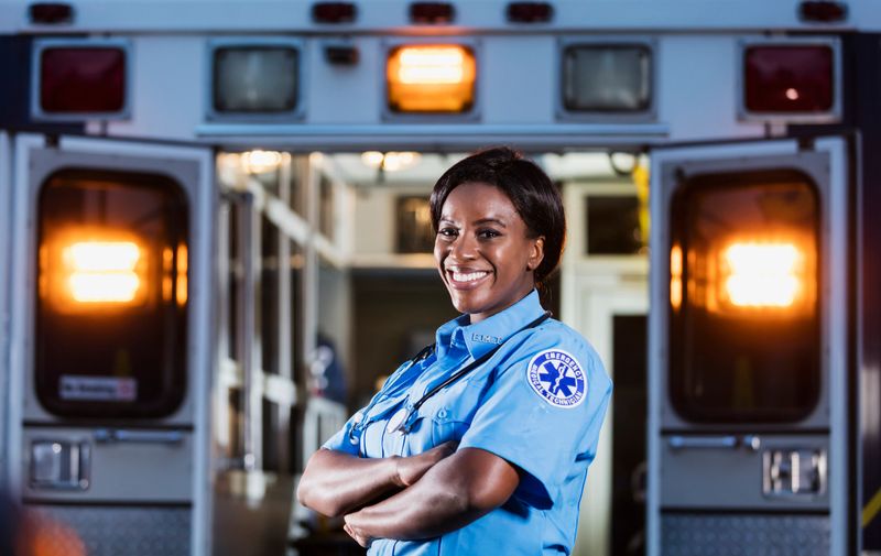 A young African-American woman standing, arms folded, smiling at the camera. The back of an ambulance with doors open and emergency lights on, is out of focus behind her. She is working as an EMT or paramedic.