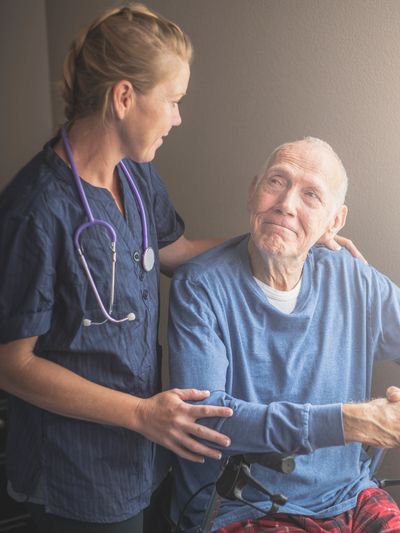 A nurse comforts an elderly man in a wheelchair near a window.