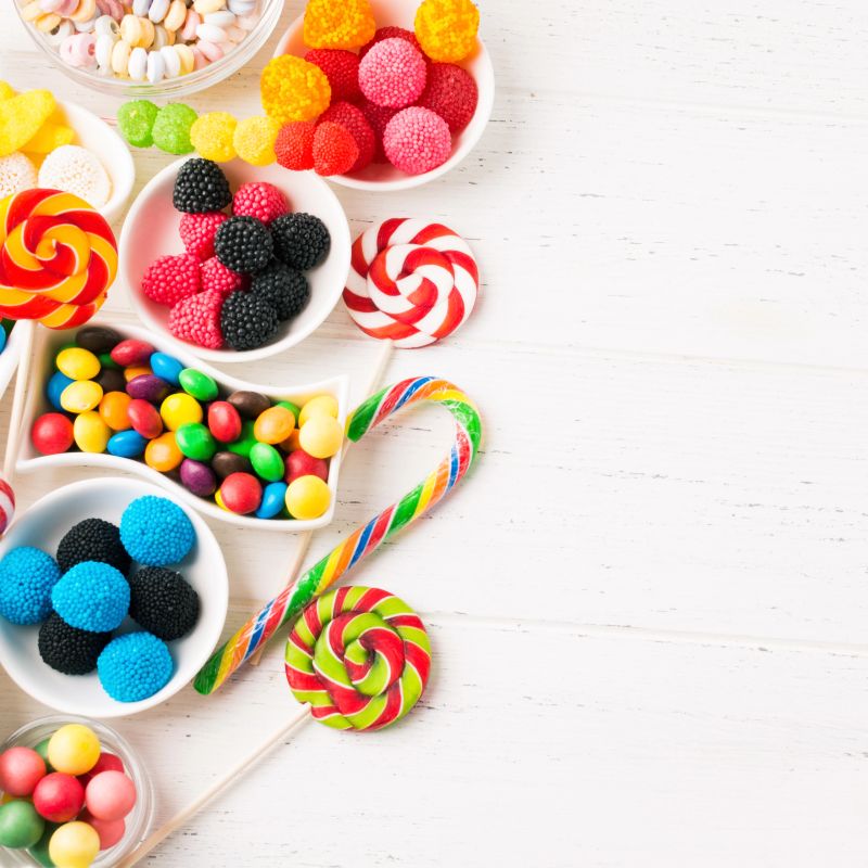 Many colorful goodies for kids in white jars over wooden table. Square shot. Flat lay. Birthday candy bar with copy space