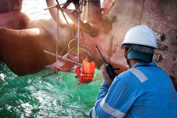 Worker in safety gear monitors a diver inspecting a ship's propeller underwater.