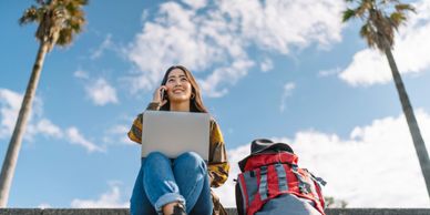 Young woman with laptop and backpack sitting on steps under blue sky.