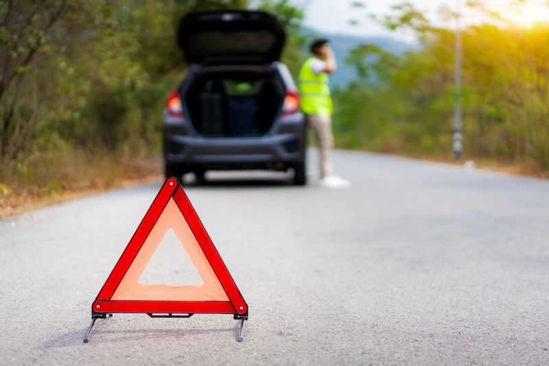 Breakdown triangle sign on road with worried Asian man talking by mobile phone with insurance or car service center company after car breakdown on country road in background.