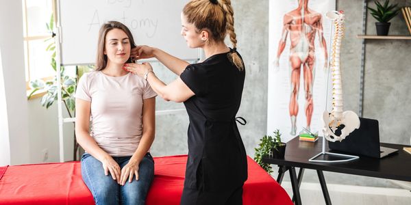 Teacher examining student's neck in anatomy class with spine model and muscle chart.