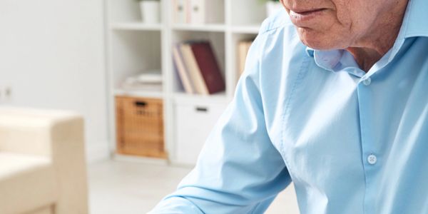Elderly man typing on a laptop at a glass table with glasses nearby.