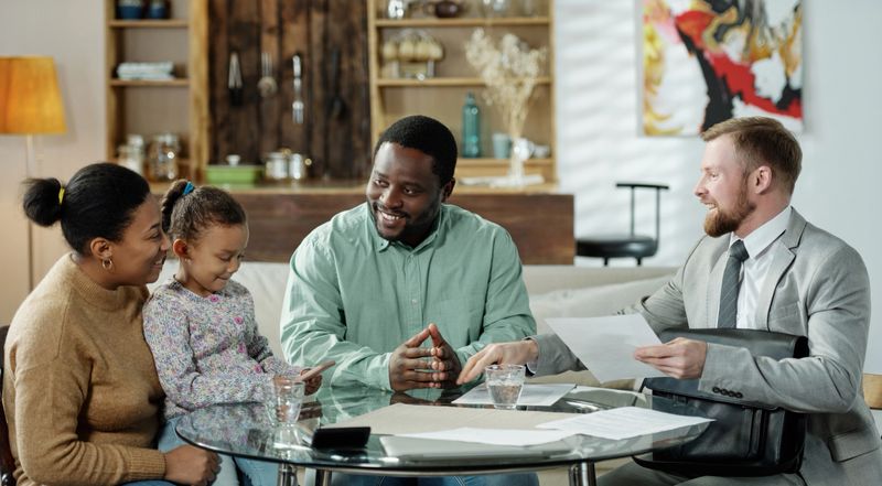 Group of cheerful ethnic couple with daughter and adult bearded agent gathering at table to discuss real estate mortgage