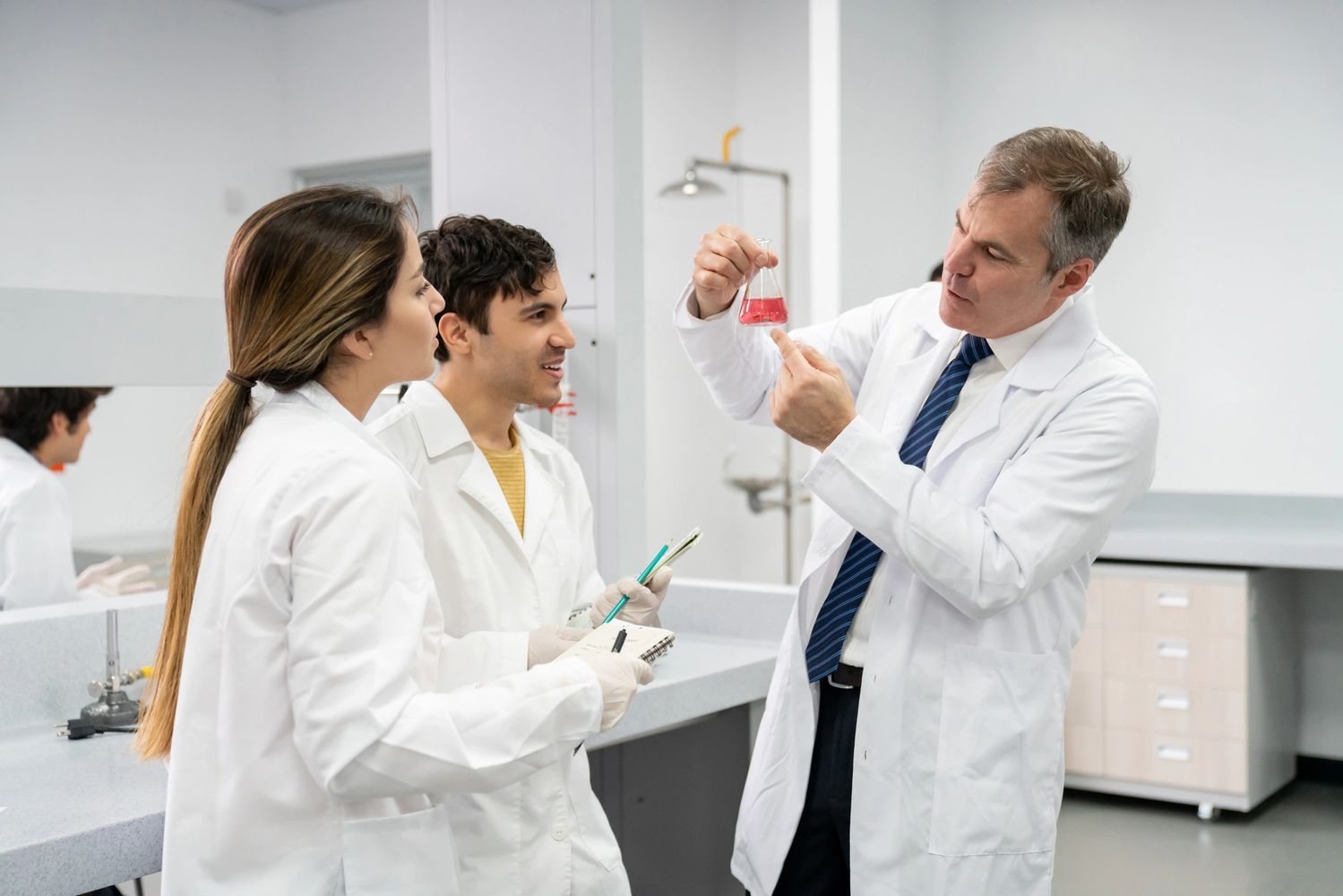 Scientist explaining a red liquid in a flask to two attentive students in a lab.