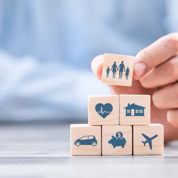 Hand stacking wooden blocks with icons of family, health, home, car, savings, and travel.