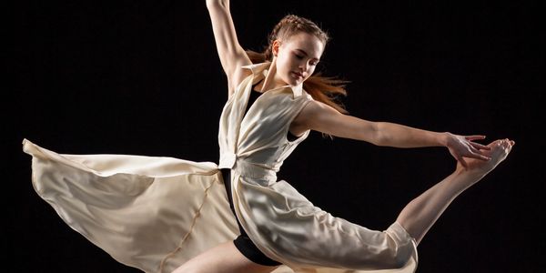 A dancer gracefully leaps mid-air in a flowing beige outfit against a black background.