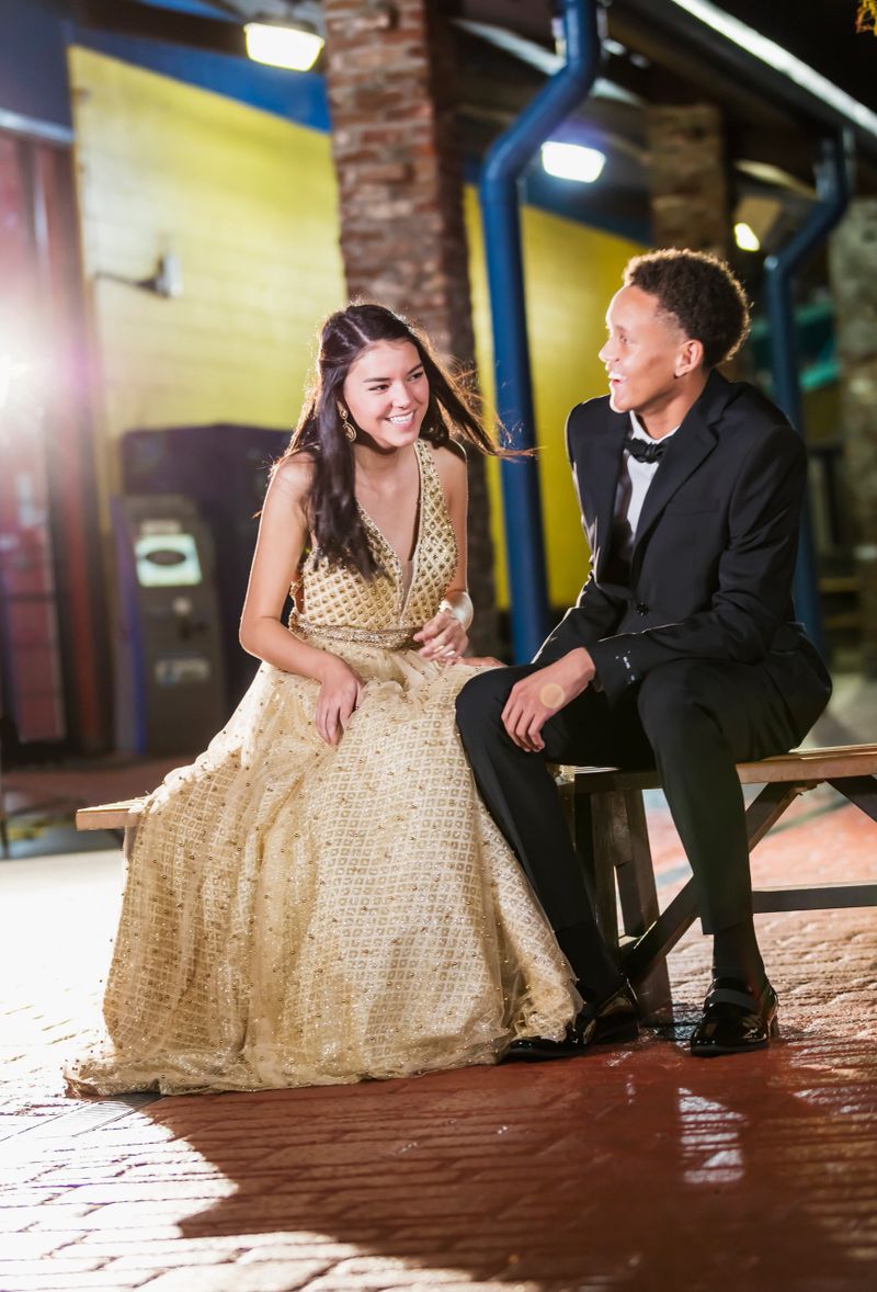 A multi-ethnic teenage couple, 17 years old, at prom, dressed in formalwear, sitting on a bench outdoors, conversing.