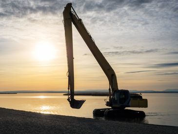 Excavator silhouetted against a sunset by the water.