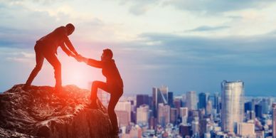 An executive in a suit helping another executive climb to the top of a mountain overlooking downtown