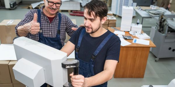 Two factory workers operate a machine, one giving a thumbs up.