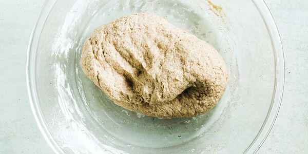 Whole wheat dough resting in a glass bowl on a white surface.