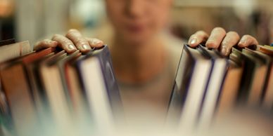 Hands browsing books on a shelf with a blurred person in the background.
