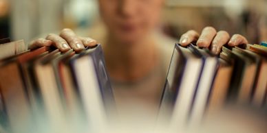 Hands browsing books on a shelf with a blurred person in the background.