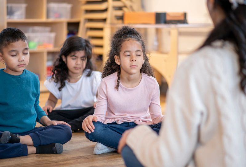A small group of multi-ethnic daycare children sit on the floor, with their legs crossed, their eyes closed and their hands on their knees.  They are each dressed casually and following their teachers verbal instructions.  The female teacher has her back to the camera and is leading the group.