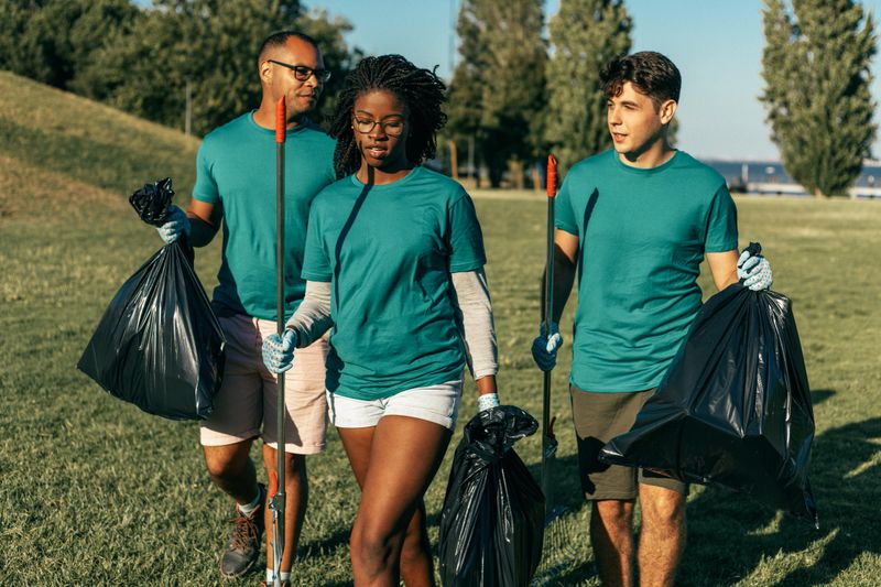 Volunteer group cleaning city park from rubbish. Young woman and men walking through lawn and carrying rakes and plastic bags. Outdoor cleaning concept