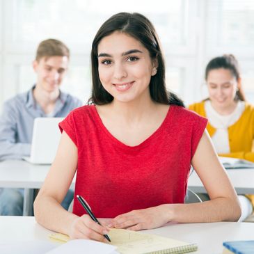 Smiling student in red shirt writing notes in classroom.